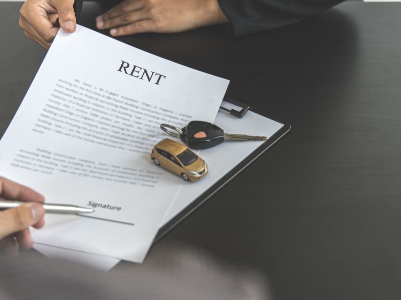 Close-up of a rental contract labeled 'RENT' on a clipboard, with a person signing and a set of car keys resting on top beside a small gold car model.